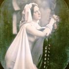 Una mujer reza arrodillada junto a una silla. Lleva un vestido de novia blanco, abierto por la parte de delante, que deja ver su pecho. Daguerrotipo estereoscópico coloreado a mano. 1850.