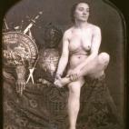 Una mujer desnuda sentada junto a un montón de armaduras. Daguerrotipo estereoscópico coloreado a mano. 1850.
