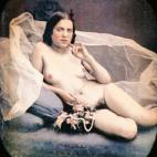 Una mujer desnuda recostada en un diván, sujetando un ramo de flores y un collar de perlas. Daguerrotipo estereoscópico coloreado a mano. 1850.
