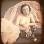 Una mujer desnuda recostada en un diván, sujetando unas flores rosas y un collar de perlas. Un velo de novia la rodea y le cubre la parte inferior del cuerpo. Daguerrotipo estereoscópico coloreado a mano. 1850.