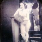 Una mujer desnuda frente a un espejo lleva zapatos, medias y una cinta en el pelo. Daguerrotipo estereoscópico coloreado a mano. 1850.