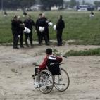 Un niño refugiado en silla de ruedas espera a que le trasladen mientras los policías antidisturbios vigilan el traslado. La policía ha asegurado que la fuerte presencia de las fuerzas antidisturbios es una mera "medida de precaución", para r...