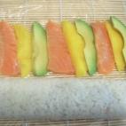 Desenvolvemos con cuidado los dos rollos de sushi y situamos el papel film encima de la mesa o tabla. Colocamos las tiras de salmón, aguacate y mango en una linea recta encima del papel film.