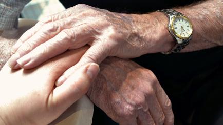 Two elderly hands holding onto one hand of a younger adult.