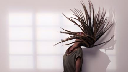 Excited stylish african woman throwing dreadlocks head back to form a hair arc, standing against wall with window shadow. 