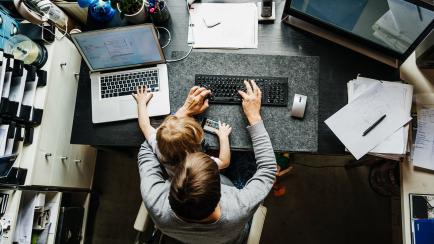 An aerial view of a mother working on a laptop in her office at hoe with her young daughter string on her lap.