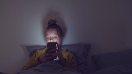 A young teenager is sitting in her bed in the dark with only the light of her phone shining on her face.