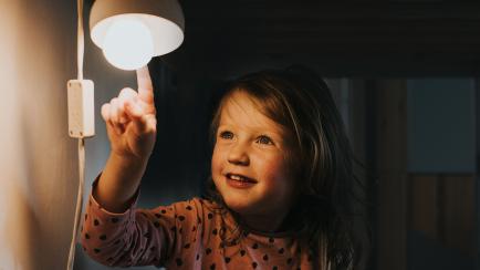 Cute young girl in pyjamas, in the dark, smiling and pointing at a little night light. Darkness provides a space for copy. Conceptual image.