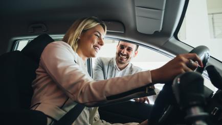 Woman buying a car in dealership sitting in her new auto, the salesman talking to her in the background