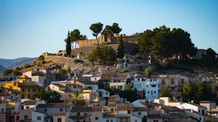 Our Lady of the Assumption Church in the town of Guadalest, the church sits on a hill with other houses. Sun setting and mountains behind.