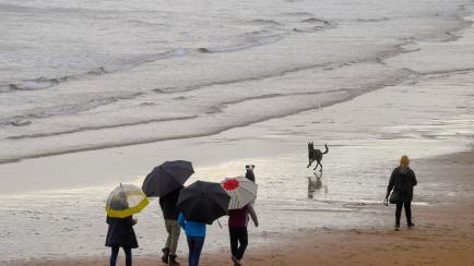 Unas personas pasean con paraguas en una playa en Gijón este sábado.