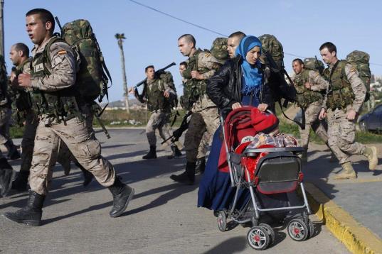 La refugiada siria Hadra Annasan empuja el carrito de su hijo Barakat en Melilla mientras varios soldados marchan para realizar unos ejercicios.
