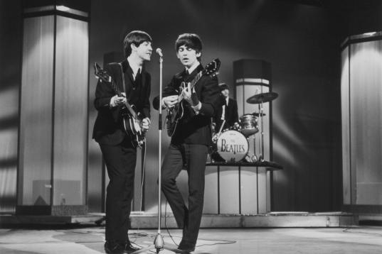 Bassist Paul McCartney and guitarist George Harrison (1943 - 2001) sing at a microphone as the Beatles perform at the London Palladium. Drummer Ringo Starr plays from a riser in the background. London, England, January 1964. (Photo by Leslie Lee...