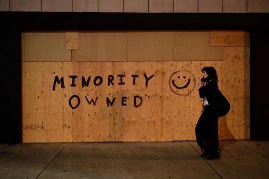 A woman walks past a store boarded up with a message as protests over the death of George Floyd continue Sunday, May 31, 2020, in Los Angeles. Armed National Guard soldiers lined the steps of Los Angeles City Hall, and cities across California d...