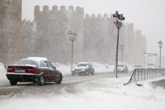 Varios vehículos circulan por una carretera próxima a la muralla de Ávila que se encuentra cubierta por la nieve caída en las últimas horas.