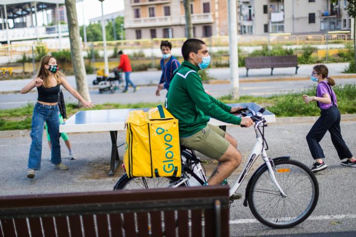 Un repartidor de Glovo circula con la bicicleta por Barcelona mientras unos niños juegan con sus padres. 