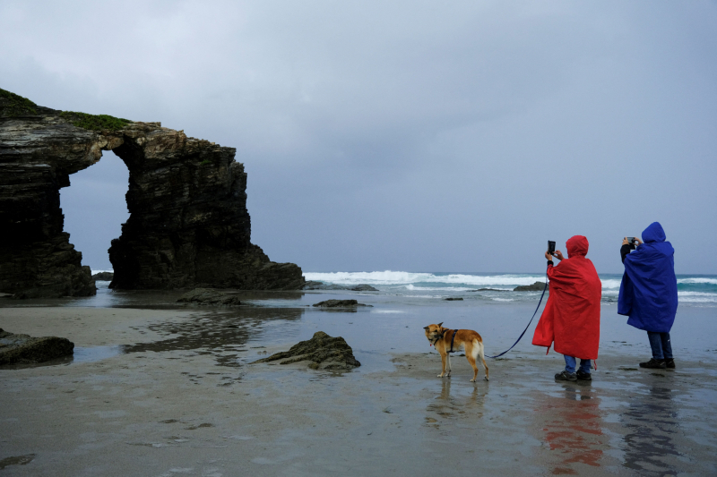 Dos turistas visitan la Praia das Catedrais cubiertos con chubasqueros.